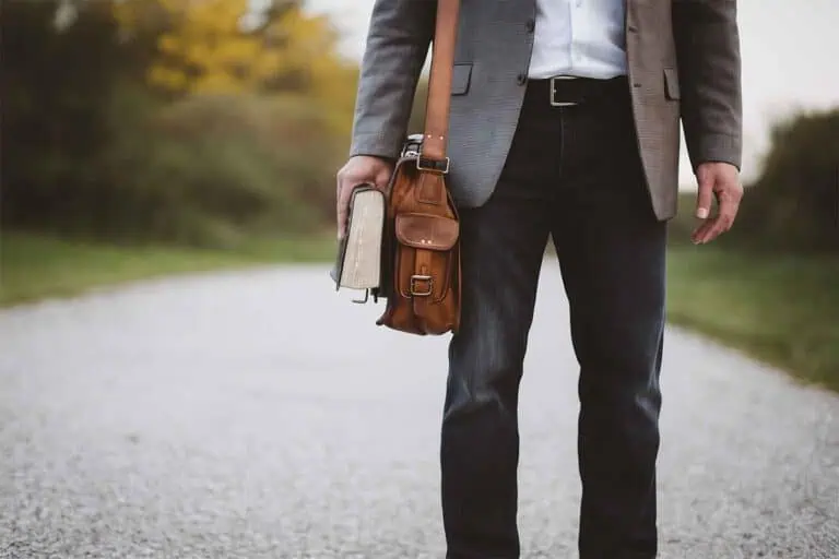 Ein Mann in Business-Kleidung steht auf einem Schotterweg, hält eine braune Ledertasche und zwei Bücher in der Hand. Der Hintergrund zeigt eine unscharfe Naturkulisse