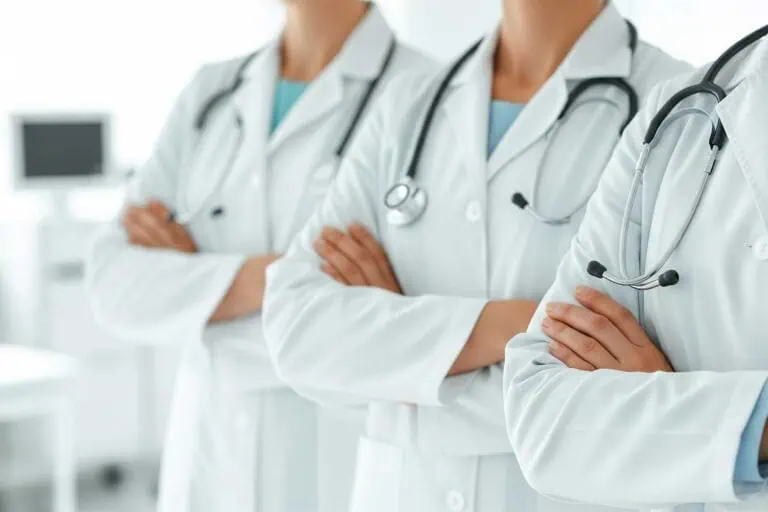 Professionelle Nahaufnahme eines dreiköpfigen Ärzteteams. Sie stehen selbstbewusst in einer Reihe mit verschränkten Armen. Alle tragen weiße Laborkittel und Stethoskope um den Hals in einer hellen, klinischen Umgebung